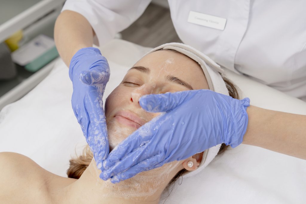 Female client receiving facial treatment with cleansing foam applied by beautician wearing blue gloves in a modern spa setting with equipment in the background